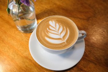 Kafe latte kahvenin yatay fotoğrafını beyaz fincanda bir yaprak tablosu, içinde çiçek olan bir cam şişe ile kapat.