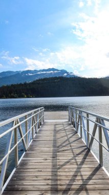 Gökkuşağı Parkı, Whistler, British Columbia, Kanada 'nın rıhtımındaki ahşap güverte rıhtımında mavi gökyüzü ve ağaçların yansıması ile güzel bir hava gününde Gökkuşağı Gölü boyunca yol ve park
