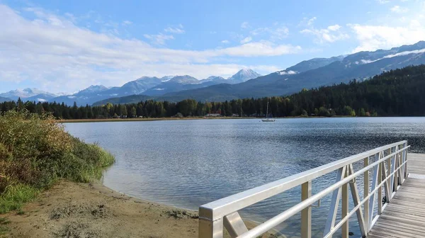 Gökkuşağı Parkı, Whistler, British Columbia, Kanada 'nın rıhtımındaki ahşap güverte rıhtımında mavi gökyüzü ve ağaçların yansıması ile güzel bir hava gününde Gökkuşağı Gölü boyunca yol ve park