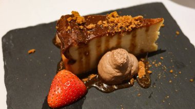 Zengin, hoşgörülü bir dilim karamelli peynirli kekin üstüne parlak çikolata sosu ve parçalanmış kurabiye parçaları. Koyu bir levhada çikolatalı mus ve taze çilekli krema.