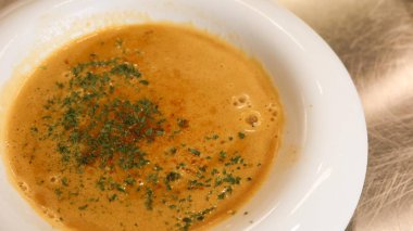 Taze yeşil otlarla süslenmiş, metalik bir yüzeyde fotokopi alanı ile servis edilen beyaz bir kâse kremalı balkabağı, turuncu-kahverengi bisk.