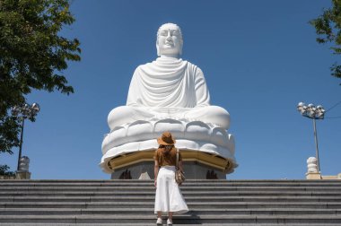 Vietnam 'ın Nha Trang kentindeki Long Son tapınağında büyük Buda ve mavi gökyüzü ile merdivenlerde duran şapkalı genç bir kadın.