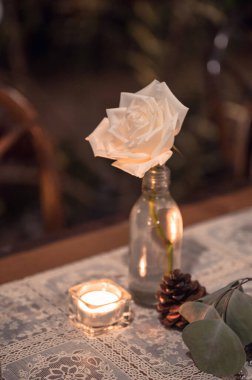 Cam şişedeki beyaz gül, mum ışığı, ahşap yemek masasında çam kozalağı.