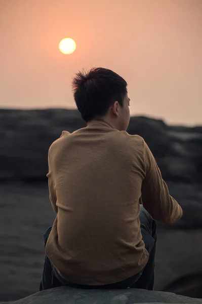 Loneliness young asian man sitting and looking at something on rock cliff at the sunset