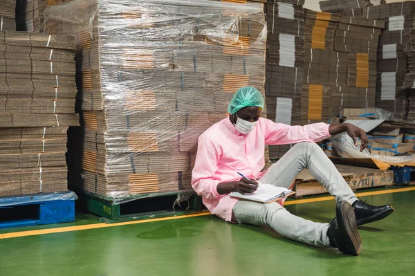 African male inventory control manager in sterile uniform sitting and checking order of cardboard box package stacked in warehouse at beverage processing plant