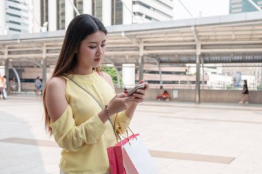 Güzel, genç Asyalı bir kadın şehirde alışveriş torbaları taşıyan akıllı telefon kullanıyor.