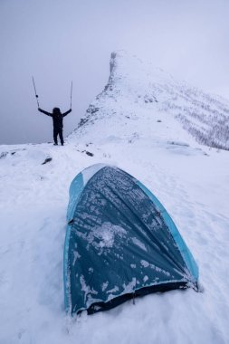 Başarılı bir dağcı, kar tepesinde çadır kurarak kar fırtınasının zirvesinde ayakta ve yürüyüş direkleri kaldırır.