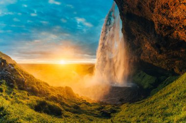 Seljalandsfoss şelalesinin yemyeşil çayırlarla ve yazın İzlanda 'nın güneyinde parlayan büyüleyici günbatımıyla nefes kesici manzarası.