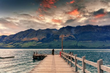 Yeni Zelanda 'da gün batımında Wakatipu Gölü ve dağa bakan Glenorchy iskelesinde durup kucaklaşan romantik çift.