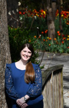 Dixon Galerisi ve Bahçeleri 'ne gelen ziyaretçi bir ağaç gövdesine ve ahşap bir köprüye dayanıyor. Arkasında laleler açıyor. Gülümsüyor ve mutlu..