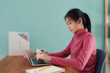 Asyalı öğrenci uzun kollu pembe elbiseler giyer. İnternetten öğrenmeyi evde bir dizüstü bilgisayardan öğrenir. Evde çevrimiçi öğrenim kavramı