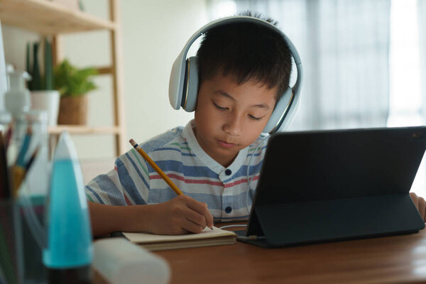 Asian boy learning online via internet with a tutor on a tablet digital with headphone, Asia child is studying while sitting in the living room at home. Concept of online learning at home 