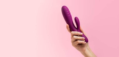 Female hand is holding purpple dildo on a pink background. Vaginal vibrator in the hand of a woman. Sex toys. Place for text. Sex shop concept. Copy space Shop for adults