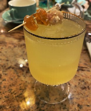 Refreshing yellow cocktail with gold rim. The drink is decorated with a wooden skewer with candied fruits, a flower, served on a marble surface. Elegant serving, evening bar or restaurant atmosphere