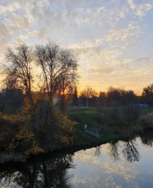 Sonbahar günbatımının ılık ışığı şehri sular altında bırakır, gökyüzünü turuncu, pembe ve altının canlı tonlarıyla boyarlar. Sakin nehir huzur ve melankolik bir güzellik manzarası yaratır..