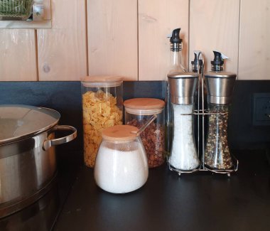 Mısır gevreği, tuz, baharat ve yağ için organik olarak tasarlanmış kavanozlarla dolu rahat bir mutfak köşesi. Ahşap kapaklı cam kaplar, metal fabrikaları ev konforu ortamı yaratır. Yemek pişirme, ev hayatı temaları