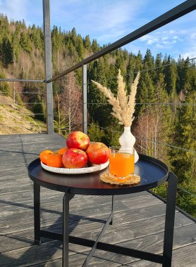 Tabaktaki meyvelerin bileşimi: muzlar, elmalar, portakallar, bir bardaktaki meyve suyu, karpatya Dağları manzaralı ahşap bir terasta siyah metal bir masada kuru bitkilerle dolu bir vazo. Görkemli zirveler