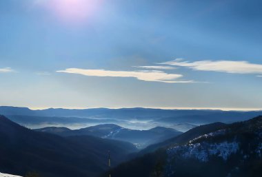 Karlı yamaçların üzerinde parlak güneşli kış manzarası. Ormanlarla kaplı dağların panoramik manzarası, vadilerde hafif sabah sisi, mavi gökyüzü ve sakin atmosfer. Seyahat için