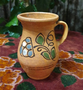 Bir kil sürahi masa örtüsüyle kaplı bir masanın üzerinde duruyor, konfor ve gelenek ortamı yaratıyor. Doğal seramik, iç mekanların özgünlüğünü, basitliğini ve sıcaklığını vurgular.