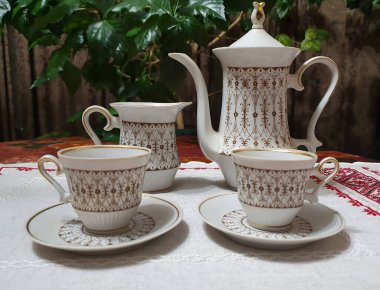 Klasik tarzda porselen kahve ve çay seti: kahve demliği, süt sürahisi ve iki fincan çay tabağı. Zarif altın sarısı süs ve yaldızlı detaylarla süslenmiş beyaz porselen işlemeli tekstil.