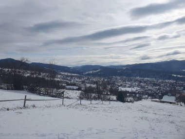 Bulutlu bir gökyüzünün altındaki vadideki bir dağ köyünün panoramik görüntüsü. Karpatların kış manzarası. Yamaçlarda evleri ve kozalaklı ormanları var.