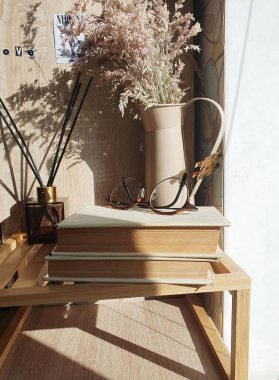 Eski kitaplar, bardaklar, vazodaki otlar ve bir sazlık ayırıcıyla rahat bir hayat. Ahşap raflı ve yumuşak gölgeli sıcak güneş ışığıyla aydınlatılmış bir iç mekan. Estetik sabah çalışma konsepti.