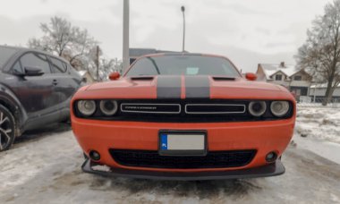 Polonya Ostrowiec Swietokrzyski 25 Ocak 2026: 11. Bulutlu bir kış gününde kırmızı Dodge Challenger otoparkta. Araba siyah yarış şeritlerine sahiptir ve karlı bir otoparkta mevzilenmiştir. Çevresinde başka araçlar ve çıplak ağaçlar vardır.