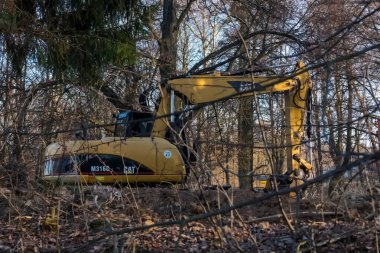 Polonya Nowa Slupia, 7 Mart 2026 saat 16: 41. Kedi, tırtıl kazıcısı ağaçların arasında inşaat alanında. Sarı bir kazıcı ormanlık bir alanda yer almaktadır. Çıplak ağaçlarla çevrili büyük kolu görülebilir. Makineler M316 modeli ile işaretlenmiş..