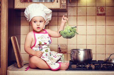 Küçük bebek bir mutfak masası ve holding brokoli üzerinde oturur