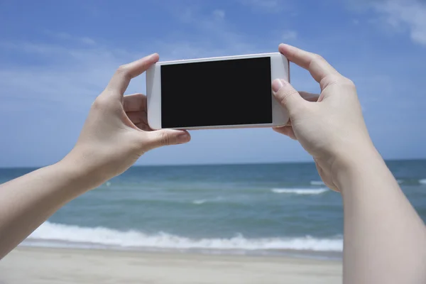 Primo piano della mano della donna che tiene smartphone, cellulare, smartphone sul mare blu sfocato bello per scattare una foto del mare, telefono con schermo nero Foto Stock