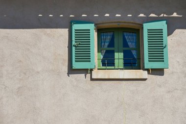 Açık panjurlu yeşil ahşap bir pencere, parlak güneş ışığında tipik bir güney Fransız mimarisi olan bej bir duvar üzerine gölgeler bırakır.