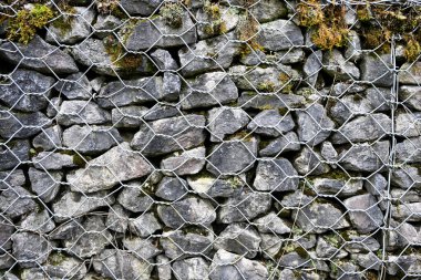 Yığılmış gri taşlar, metal bir tel örgüyle bir arada tutularak erozyon kontrolü ve peyzaj için sağlam bir Gabion duvarı oluşturur.
