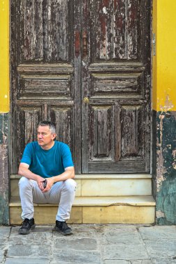 Bir adam, Sevilla 'da sarıya boyanmış eski ahşap bir kapının önünde düşünceli bir şekilde oturur, eskimiş dokular ve zamansız kentsel bir anda sessizce tefekkür eder.