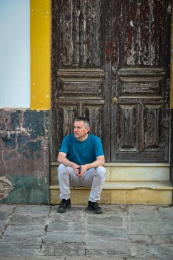 Bir adam, Sevilla 'da sarıya boyanmış eski ahşap bir kapının önünde düşünceli bir şekilde oturur, eskimiş dokular ve zamansız kentsel bir anda sessizce tefekkür eder.