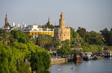 Sevilla 'daki Guadalquivir Nehri' nin yanındaki Torre del Oro manzarası, palmiye ağaçları, botlar ve tarihi mimariyle çevrili. Altın kule öğleden sonra ışığında parlıyor.