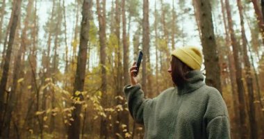 Sarı bereli ve yün ceketli mutlu bir genç kadının güzel bir sonbahar ormanında selfie çekerken ya da kendi videosunu çekerken orta boy bir resmi. Arka plan bir...