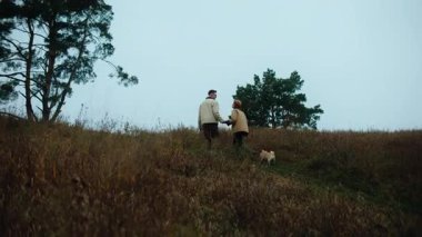 Geniş açı, mutlu genç bir çiftin geniş, manzaralı bir vadi ya da tepe manzarasında yürürken el ele tutuşmalarını takip eden geniş açılı bir çekim. Köpekleri patikanın önünde koşuyor.