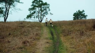 Geniş açı, mutlu genç bir çiftin geniş, manzaralı bir vadi ya da tepe manzarasında yürürken el ele tutuşmalarını takip eden geniş açılı bir çekim. Köpekleri patikanın önünde koşuyor.
