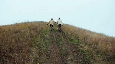 Dinamik bir el kamerası görüntüsü, mutlu ve neşeli genç bir çifti, sonbahar manzarasında çimenli bir tepeye çıkıp el ele tutuşup gülerken takip eder. Yüzleri görünüyor. Çekimin sonunda...