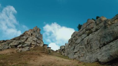 Statik geniş açı çarpıcı bir dağ manzarasını yakalar. Büyük, dramatik kaya oluşumları ve kayalıklar çimenli bir tepeden yükseliyor. Kayaların arasında küçük, uzak bir taş ya da ahşap bir kulübe var.