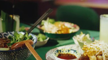 Restoran masasında yeşil bir kasede servis edilen geleneksel Thai Pad Thai eriştesi. Tabak fıstıklı fasulye filizi ve limonla süslenmiş. Bulanık ön planda...