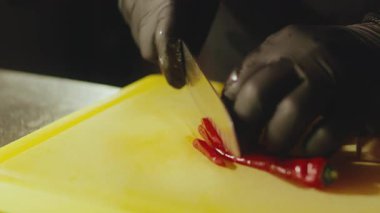 Profesyonel aşçı ellerinin siyah koruyucu eldivenler giyip sarı plastik bir kesme tahtasına keskin bir bıçakla taze kırmızı acı biber keserken yakın çekim görüntüsü. Baharatlı malzemelerin hazırlanması