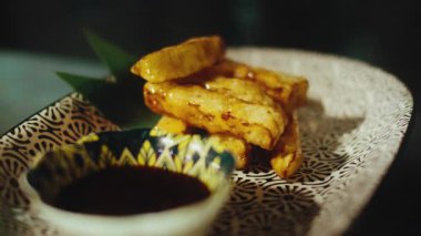 Profesyonel şefin, tatlı ve ekşi sosla kaplanmış kızarmış patlıcan çubuklarını düzenlemek için metal cımbız kullanırken yakın plan çekimi. Aperatif güzel desenli seramik tabakta servis ediliyor.