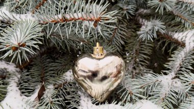 Huzurlu bir kış atmosferinde, karla kaplı köknar dalında parıldayan kalp şeklinde bir süs.