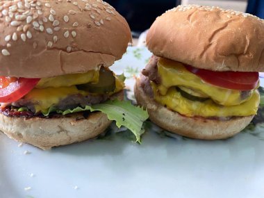 Two homemade cheeseburgers with melted cheese, lettuce, tomatoes, and cucumbers on a white plate
