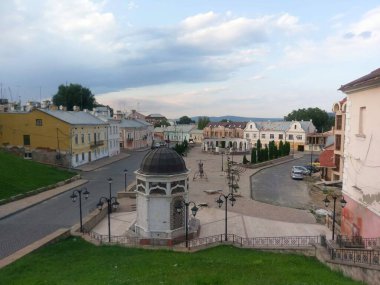 Bulutlu bir gökyüzü altında çeşitli stillerin pavyonu, heykelleri ve mimarisi olan sakin bir şehir meydanı.