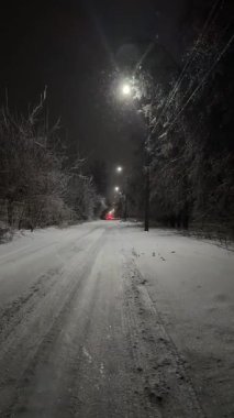 Kar yağışı, sokak lambaları, buzlu ağaçlar ve elektrik hatlarıyla kaplı bir gece yolu atmosferik kış manzarası.