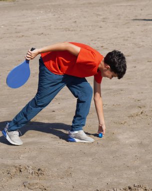 Genç bir çocuk kumsalda tenis oynarken topu topluyor.