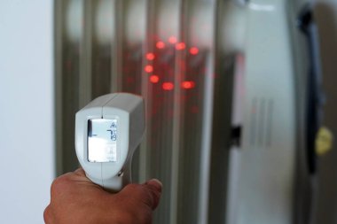 Beyaz bir radyatörün ısısını ve doğru işlevini ölçen temassız dijital kızılötesi bir termometreyle el.