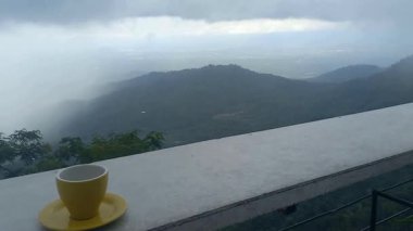 Yüksek irtifada, dağlar ve sisli gökyüzü manzaralı bir restoranda bir fincan sıcak içeceğin tadını çıkar. Tumpeng Menoreh, Yogyakarta, Endonezya 'da yer alan bu yer harika bir doğal turizm beldesi..